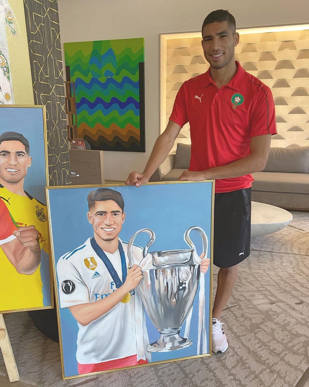Achraf Hakimi standing beside his framed portrait painting, holding a Champions League trophy, in a hotel lounge.