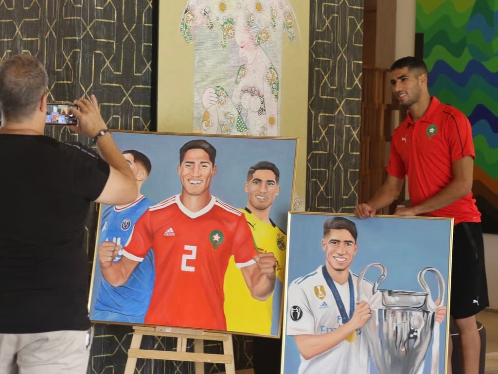 A man photographing three framed footballer portraits on an easel, including Achraf Hakimi in Morocco's red jersey.