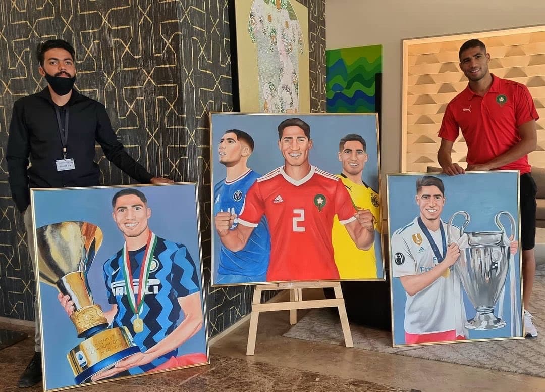 A hotel staff member and Achraf Hakimi posing beside two framed portrait paintings.