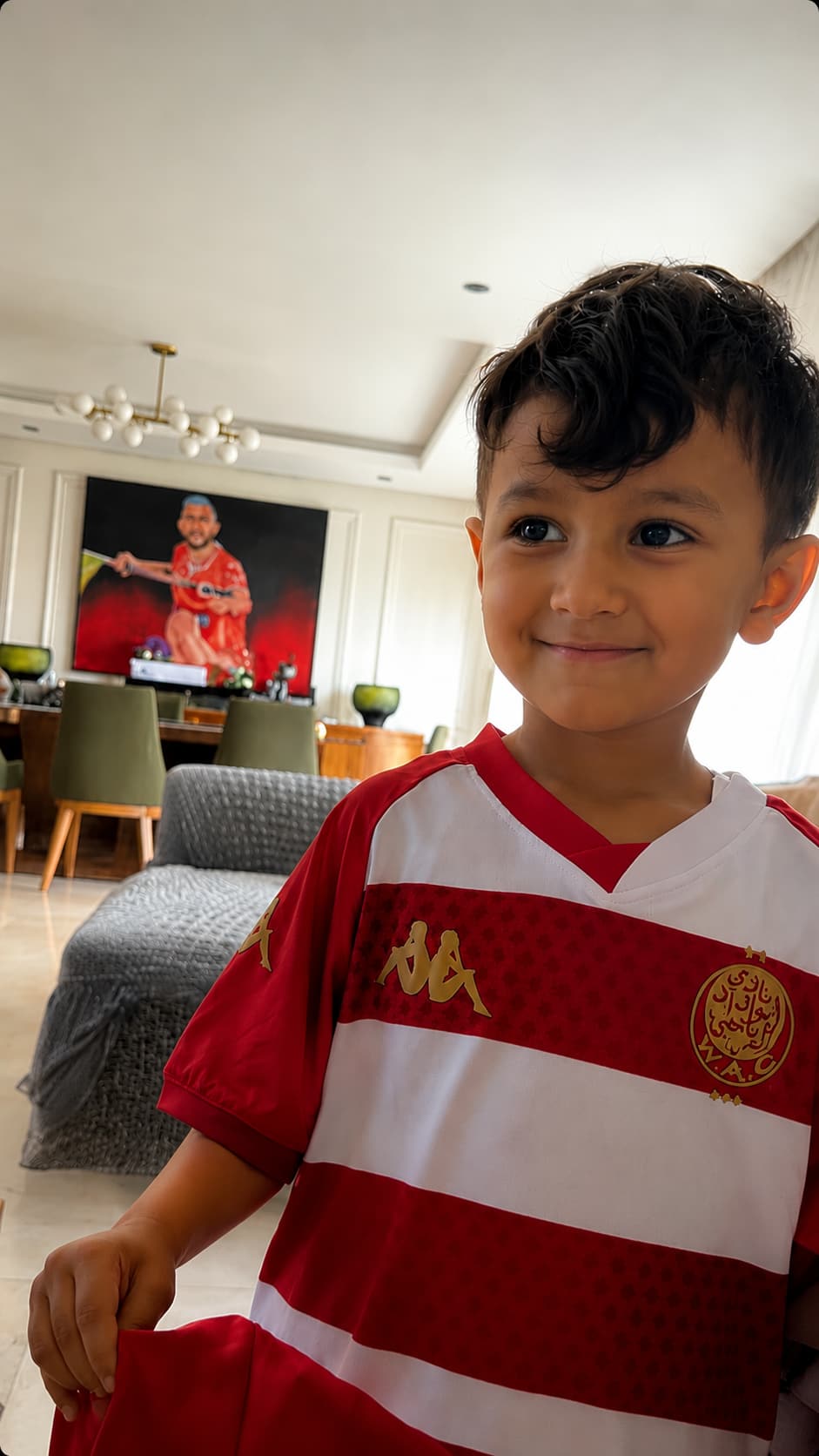 A young boy in a Wydad Athletic Club jersey standing in front of the framed portrait painting.