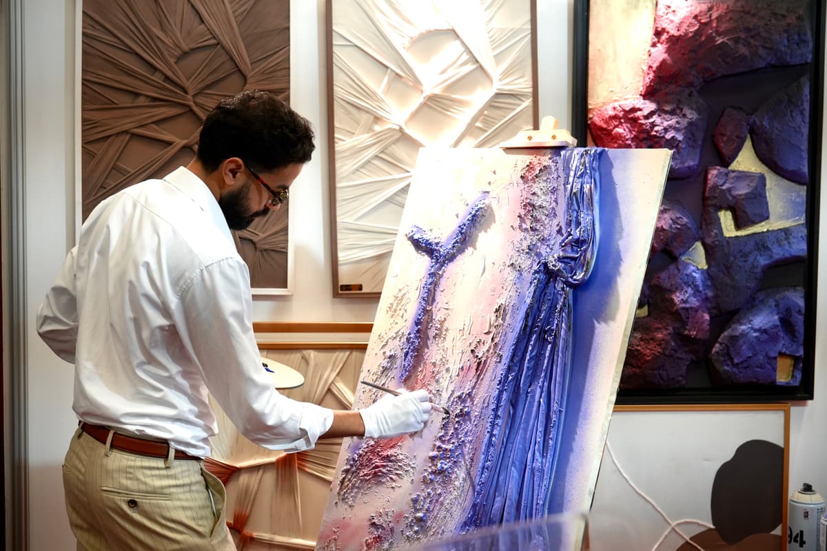 Kamal Hannouda applying gold pigment to a textured abstract canvas at his easel, surrounded by other textured artworks in the studio.