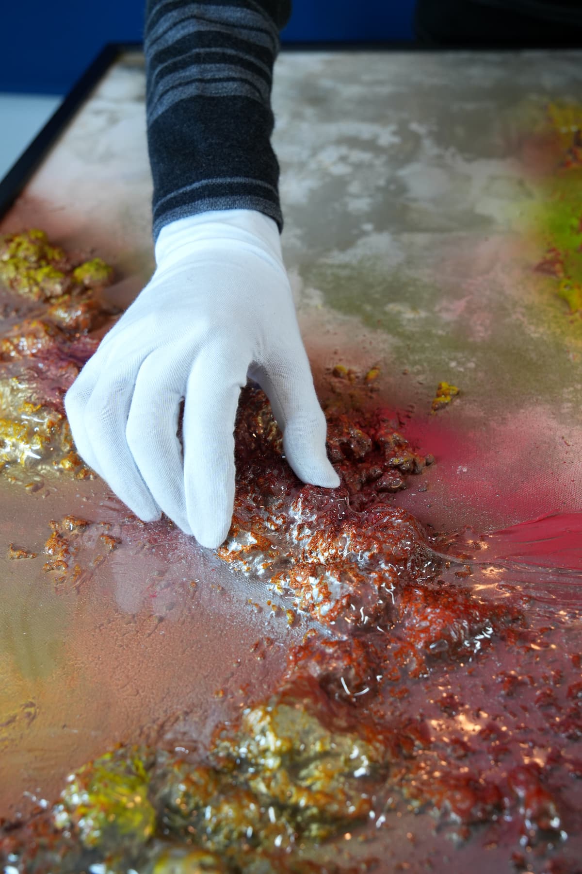 Close-up of a gloved hand applying gold and red pigment to a richly textured abstract canvas.