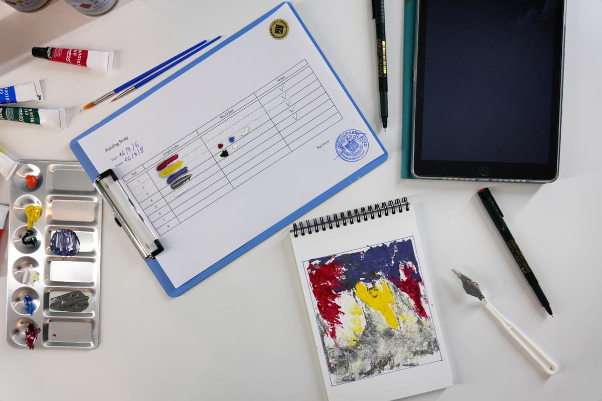 A flat-lay of studio materials: oil paint tubes, brushes, a Calligraphie Joud painting-study sheet, and a small color-study sketch.