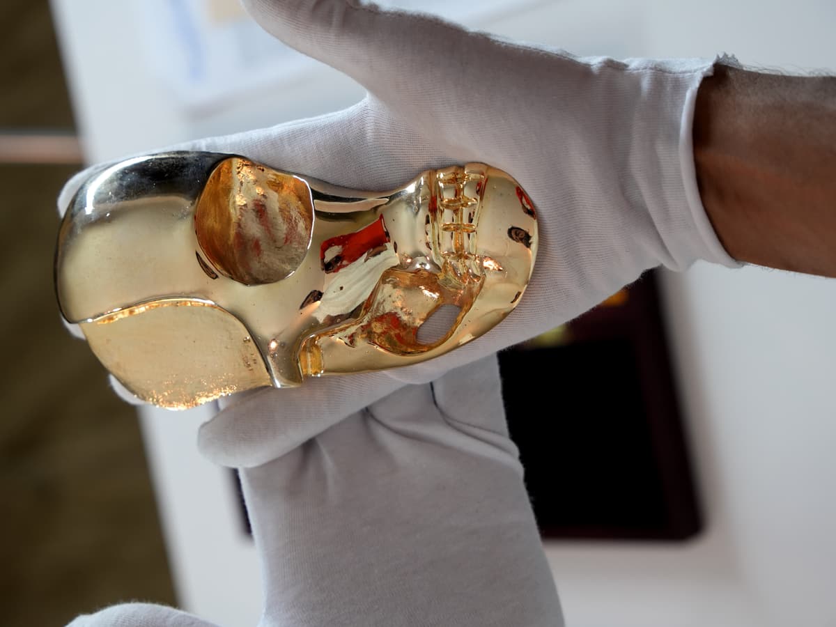 Close-up of a gloved hand holding a polished gold sculpture shaped like a stylized half-skull.