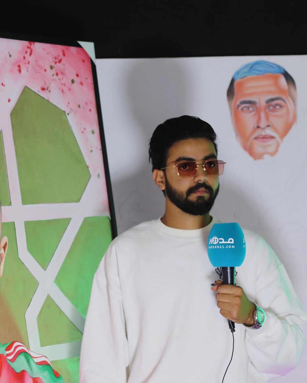 Kamal Hannouda being interviewed on camera, holding a branded microphone, beside a football-themed painting.