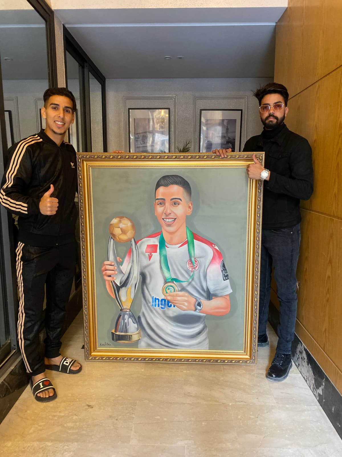 A footballer smiling beside his framed portrait painting, which shows him holding a trophy and medal.