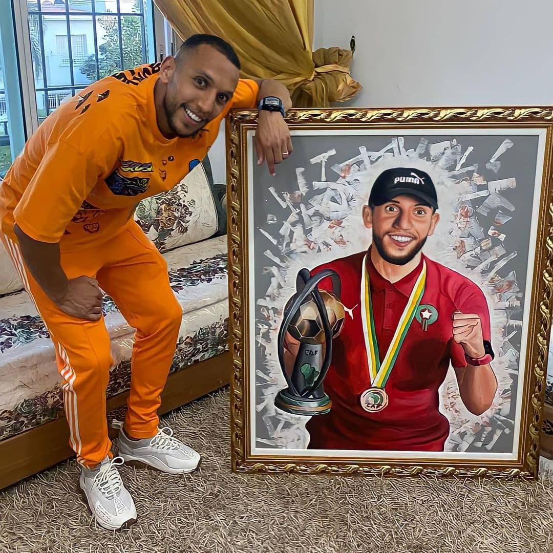 Two men holding a framed portrait painting of a footballer in a hallway.